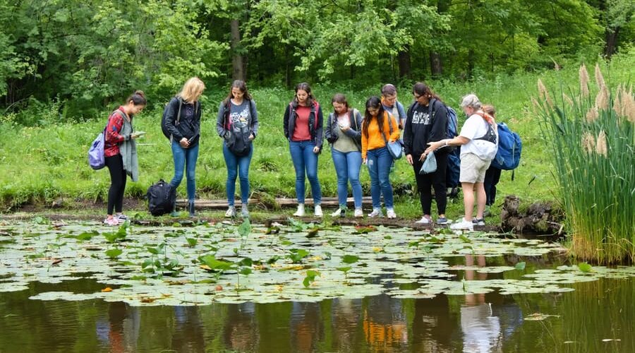 A diverse group of local residents and a wildlife expert collaboratively studying natural deterrent methods and animal tracks around a pond. The scene highlights community involvement in pond predator management.