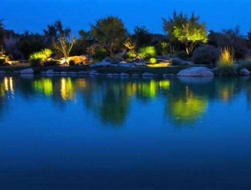Evening pond illuminated with a mix of blue and green lights creating depth, and amber and red tones highlighting surface features, reflecting a magical interplay of colors on the water.