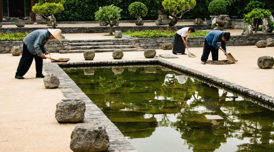 "Men constructing stonework and women tending plants in a traditional water garden setting, illustrating historical gender roles."