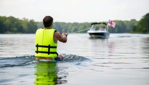 A water skier wearing a bright life jacket is being towed on a private pond, with a safety flag and boat in the background.